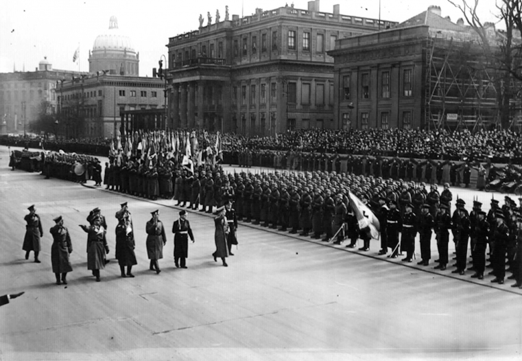 Hitler, accompanied by Grand Admiral Raeder, Generals Keitel and Milch, Reichsführer-SS Himmler, Lieutenant General Hase, General Reinhardt, and the Reichskriegsopferfuhrer Oberlindober, marches in front of the Ehrenbataillon. 